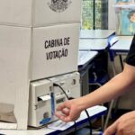 Mesário da Justiça Eleitoral ao lado de urna eletrônica dentro da cabina de votação