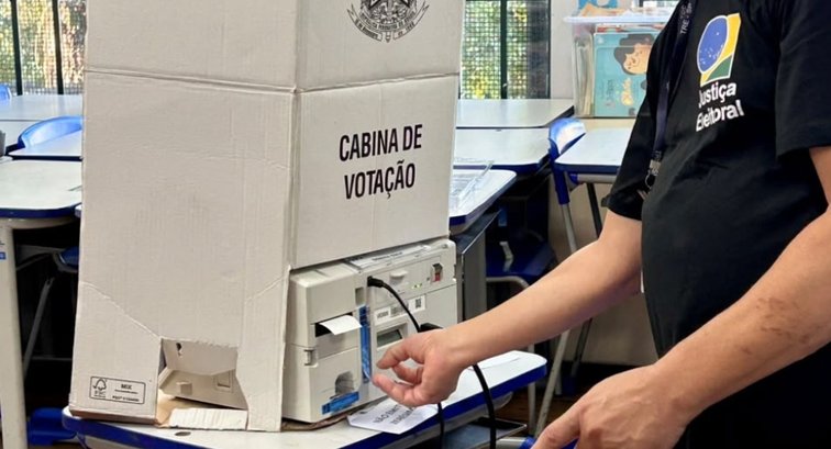 Mesário da Justiça Eleitoral ao lado de urna eletrônica dentro da cabina de votação